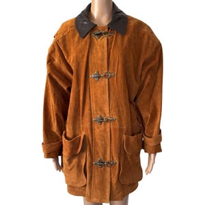 May include: A long-sleeved, tan suede jacket featuring a dark brown leather collar, two large front patch pockets, and four metallic toggle closures down the center front. The jacket has a relaxed fit with structured shoulders and long sleeves.