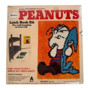 May include: A vintage Peanuts latch hook kit featuring Snoopy holding a blue object against an orange background. The kit includes a full-color design on cotton canvas and pre-cut acrylic yarn. The finished size is approximately 20 x 27 inches.