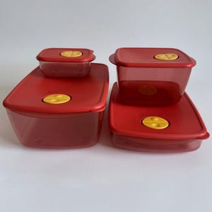 Op de afbeelding: Een set van vier doorschijnende rode plastic voedselbewaarbakjes met bijpassende rode deksels. Elk deksel is voorzien van een ronde gele ontluchtingsdop. De bakjes variëren in grootte en omvatten zowel rechthoekige als vierkante vormen.