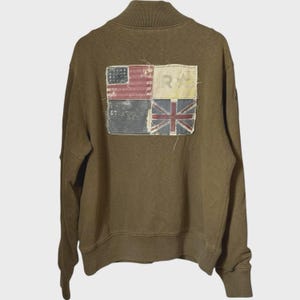 May include: An olive green long-sleeve sweatshirt featuring a ribbed collar and cuffs. The back displays a large, rectangular, frayed-edge patch divided into four quadrants, showcasing various flag-inspired designs, including a star and a Union Jack.