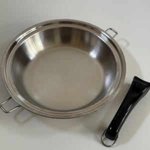 Puede incluir: Una sartén redonda de acero inoxidable con interior pulido y dos pequeñas asas laterales de metal. Un mango de plástico negro desmontable con una anilla metálica en el extremo se encuentra colocado por separado junto a la sartén sobre una superficie.