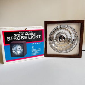 May include: A Realistic Wide Angle Strobe Light, displayed in a wooden frame and its original box. The strobe light has a clear, faceted lens. The box is white with a red and blue design and the text "Realistic Wide Angle Strobe Light".