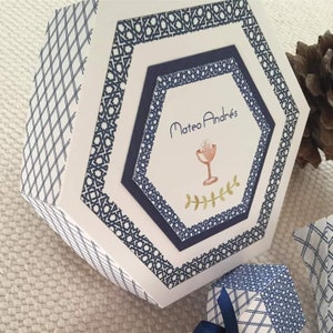 Peut inclure: Une boîte hexagonale blanche avec un motif géométrique bleu et blanc. La boîte a une petite fenêtre hexagonale avec une bordure bleue et le texte "Mateo Andrés" à l'intérieur. La fenêtre présente également une petite illustration d'un calice.