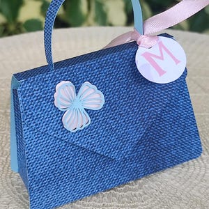 Puede incluir: Un bolso de mano azul con estampado de mezclilla, con una cinta rosa y una etiqueta circular blanca con la letra "M" en rosa. El bolso de mano tiene un asa azul y un recorte de mariposa azul en la parte delantera.