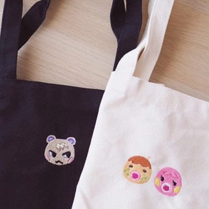 May include: Two tote bags, one black and one white, with embroidered character designs. The black bag features a grumpy bear face, while the white bag displays two round-faced characters. Both bags have long straps.