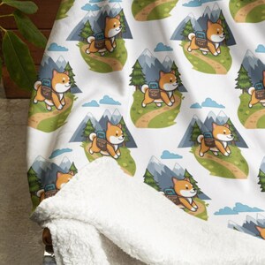 May include: White blanket with a repeating pattern of a Shiba Inu dog with a backpack hiking in the mountains. The blanket has a soft, white sherpa lining. The design includes green trees, blue mountains, and clouds.