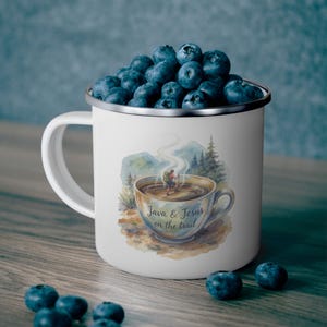 Puede incluir: Taza de camping de esmalte blanco llena de arándanos. La taza presenta un diseño de acuarela de una taza de café con un excursionista y el texto "Java & Jesus on the trail". La taza tiene un borde plateado y un asa blanca.