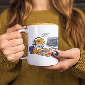 May include: White ceramic mug featuring a cartoon bearded dragon wearing a headset and working on a computer. The mug has the text "DID YOU TRY TURNING IT OFF, AND ON AGAIN." A heart is on the computer screen.