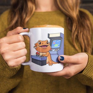 May include: White ceramic mug featuring a cartoon bearded dragon playing an arcade game. The dragon is orange and brown, with a happy expression. The arcade cabinet is blue and gray, with a dinosaur graphic on the screen. The mug has a white handle.