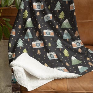May include: A black throw blanket with a repeating pattern of cameras, trees, and stars. The cameras are orange and white, and the trees are green and blue. The blanket has a white, fluffy underside.