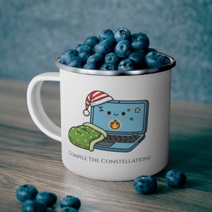 May include: White enamel mug filled with blueberries, featuring a cartoon laptop wearing a Santa hat and a green sleeping bag. The mug has the text "COMPILE THE CONSTELLATIONS" on it.