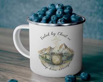 Peut inclure: Mug en émail blanc rempli de myrtilles, avec un motif aquarelle d'une tasse à café, de montagnes et d'une croix. Le texte indique "Fueled by Christ and camp-brew courage". La tasse a un bord argenté et une anse blanche.