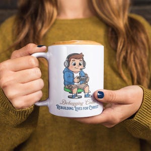 May include: White ceramic mug featuring a cartoon illustration of a boy with headphones and a game controller. The boy is wearing a blue jacket and shorts. The mug has the text "Debugging Code, Rebuilding Lives for Christ."