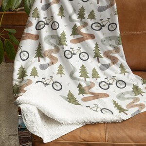 May include: White throw blanket with a repeating pattern of bicycles, trees, and winding trails in shades of brown, green, and gray. The blanket has a soft, white sherpa lining. The blanket is draped over a brown leather couch.