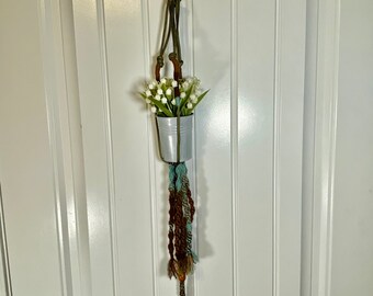 May include: A macrame plant hanger with a white flower pot and white flowers hanging from a white door. The hanger is made of brown and blue yarn.