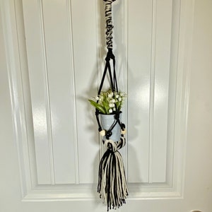 May include: A black and white macrame plant hanger with a wooden ring at the top. The hanger is holding a small metal pot with white flowers.