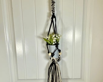 May include: A black and white macrame plant hanger with a wooden ring at the top. The hanger is holding a small metal pot with white flowers.
