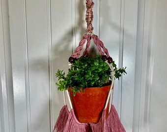 May include: A pink macrame plant hanger with wooden beads and tassels, holding a terracotta pot with a green plant.