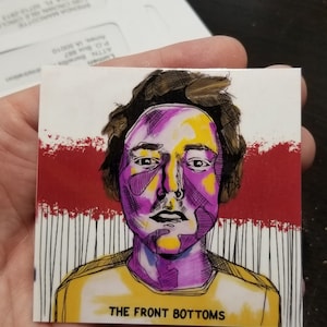 May include: A square art print featuring a stylized portrait with yellow, purple, and black ink. The subject has brown hair and is wearing a yellow shirt. The text "THE FRONT BOTTOMS" is printed at the bottom.