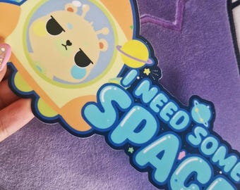 Op de afbeelding: Een gele en oranje raket met een cartoon-alien erin. De raket vliegt door de ruimte met sterren en planeten. De tekst "Ik heb wat ruimte nodig" staat in blauwe letters.