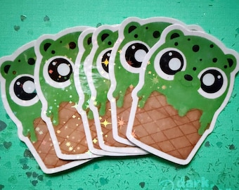 Op de afbeelding: Een verzameling stickers met een groen ijsje in de vorm van een beer. Het ijs heeft een druppeleffect en de wafel is bruin. De stickers hebben een witte rand en zijn gerangschikt op een groene ondergrond met glitter.