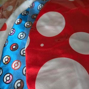 Puede incluir: Tres cuadrados de tela con diferentes patrones. El primer cuadrado es azul con lunares blancos y un patrón de Poké Balls rojas, blancas y azules. El segundo cuadrado es blanco con lunares rojos. El tercer cuadrado es rojo con lunares blancos.