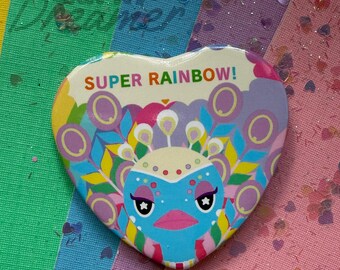 Op de afbeelding: Hartvormige button met een kleurrijk pauwontwerp en de tekst "SUPER RAINBOW!". De button heeft een blauwe pauw met stervormige ogen, omringd door paarse cirkels en kleurrijke veren. De achtergrond is een regenboog van kleuren.
