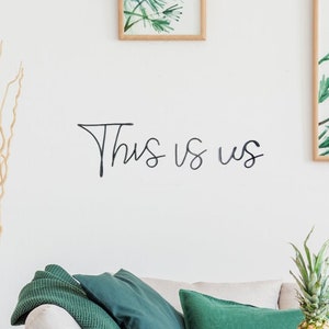 Puede incluir: Una pared blanca con las palabras "This is us" escritas en cursiva negra. Hay un sofá con almohadas verdes y una piña sobre una mesa de madera delante del sofá.