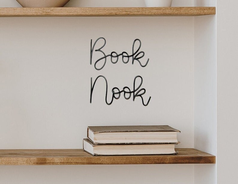 Puede incluir: Una pared blanca con un letrero de metal negro que dice "Book Nook". Dos libros est&aacute;n apilados en un estante de madera debajo del letrero.