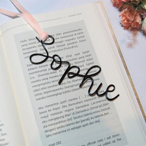 May include: A black wire bookmark shaped like the word "Sophie" is shown on top of an open book. The bookmark is resting on a page with text in a foreign language.