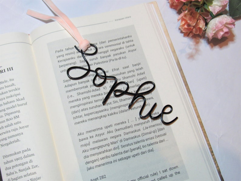 May include: A black wire bookmark shaped like the word "Sophie" is shown on top of an open book. The bookmark is resting on a page with text in a foreign language.