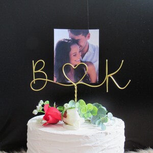 Könnte beinhalten: Ein Tortenaufsatz für eine Hochzeit mit einem Foto eines Paares, goldenen Drahtinitialen "B & K" und einem Herz. Der Kuchen ist mit einer roten Rose, einer weißen Rose und grünem Laub geschmückt. Der Hintergrund ist schwarz.