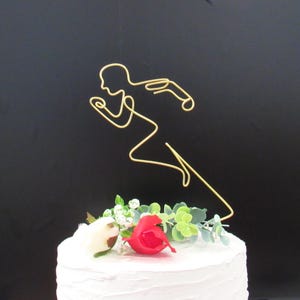 May include: A gold wire runner cake topper. The topper is placed on a white frosted cake decorated with red and white flowers and greenery. The background is black.