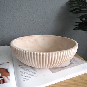 May include: A light pink ceramic bowl with a ribbed texture. The bowl is sitting on a white and grey book.