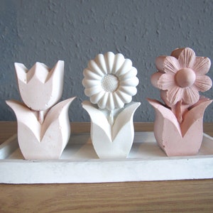 K&ouml;nnte beinhalten: Drei dekorative Blumenskulpturen auf einem wei&szlig;en Tablett. Die Blumen sind aus wei&szlig;em und rosa Gips gefertigt und in einer Reihe angeordnet. Die Blumen sind eine Tulpe, eine G&auml;nsebl&uuml;mchen und eine Blume mit f&uuml;nf Bl&uuml;tenbl&auml;ttern.