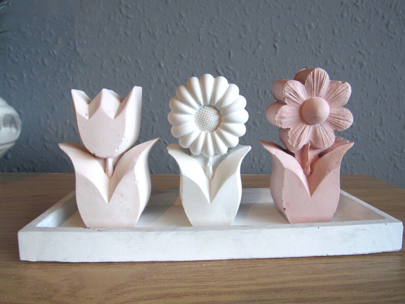 K&ouml;nnte beinhalten: Drei dekorative Blumenskulpturen auf einem wei&szlig;en Tablett. Die Blumen sind aus wei&szlig;em und rosa Gips gefertigt und in einer Reihe angeordnet. Die Blumen sind eine Tulpe, eine G&auml;nsebl&uuml;mchen und eine Blume mit f&uuml;nf Bl&uuml;tenbl&auml;ttern.