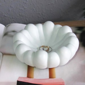 Puede incluir: Plato para anillos de cerámica blanca con un borde rizado, con forma de flor. Dos anillos de oro descansan en el centro del plato.