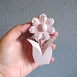 K&ouml;nnte beinhalten: Eine rosafarbene Keramik-Blumenskulptur mit einer einzelnen Bl&uuml;te und Bl&auml;ttern. Die Bl&uuml;te hat eine runde Mitte und f&uuml;nf Bl&uuml;tenbl&auml;tter.