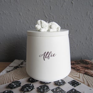 May include: White ceramic pet urn with a small white dog figurine on the lid. The urn is inscribed with the name "Alfie".