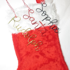 Puede incluir: Un calcetín navideño rojo con un borde de piel sintética blanca, adornado con etiquetas de nombre de alambre. Las etiquetas deletrean "Rudolph", "Santa" y "Sophie" en alambre dorado, rojo y negro, con colgadores de hilo rojo y blanco.