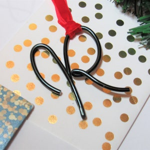 May include: Black metal wire ornament shaped like the letter R, with a red ribbon attached. The ornament is on a white background with gold polka dots.