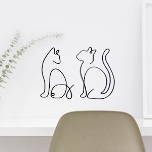 May include: Two black wire sculptures of cats, one with a fish, on a white wall.