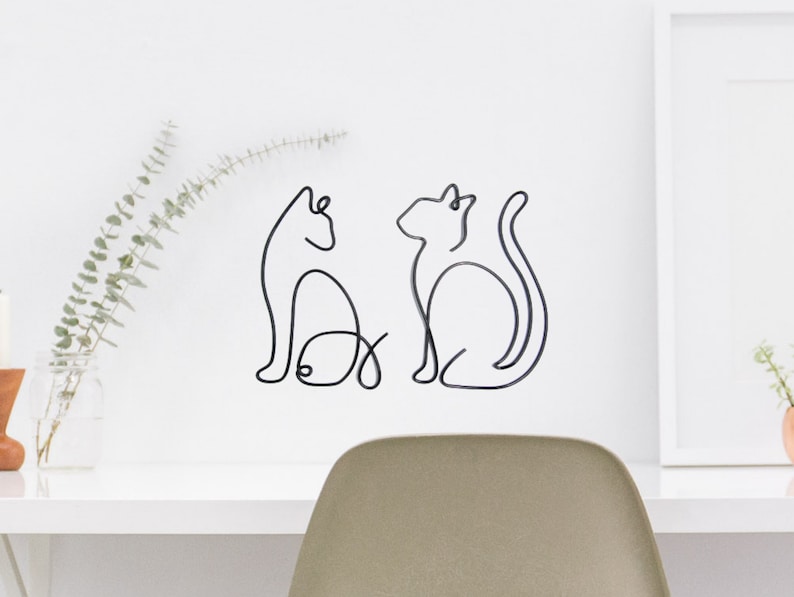 May include: Two black wire sculptures of cats, one with a fish, on a white wall.