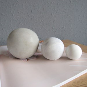 May include: Three smooth, round concrete spheres in various sizes. The spheres are light grey and white in colour and are arranged in a row on a pink and white magazine.