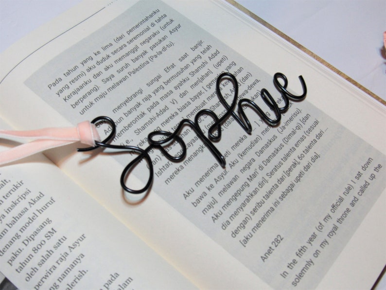 May include: A black wire bookmark shaped like the word "Sophie". The bookmark is laying on a page of an open book. The book is white with black text.