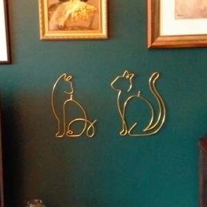May include: Two gold metal cat silhouettes on a teal green wall. The cats are facing different directions and are in a simple line art style.