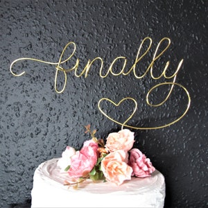 May include: Gold wire cake topper with the word "finally" and a heart. The topper is on a white cake with pink flowers.
