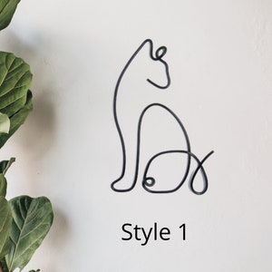 Op de afbeelding: Een zwart draadbeeld van een kat die zit met zijn staart om zijn lichaam gekruld. Het beeld is op een witte muur met de tekst "Style 1" eronder.
