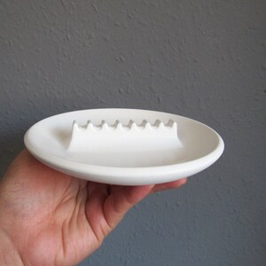 May include: White ceramic ashtray with a wavy, indented line for holding cigarettes.