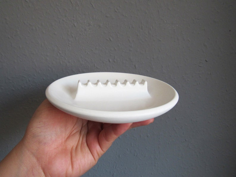 May include: White ceramic ashtray with a wavy, indented line for holding cigarettes.